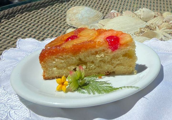 Em um prato de louça branco e raso está uma fatia de bolo de abacaxi com cerejas por cima e enfeitado com uma flor também em cima do prato. Ao fundo, conchas do mar decoram a mesa.
