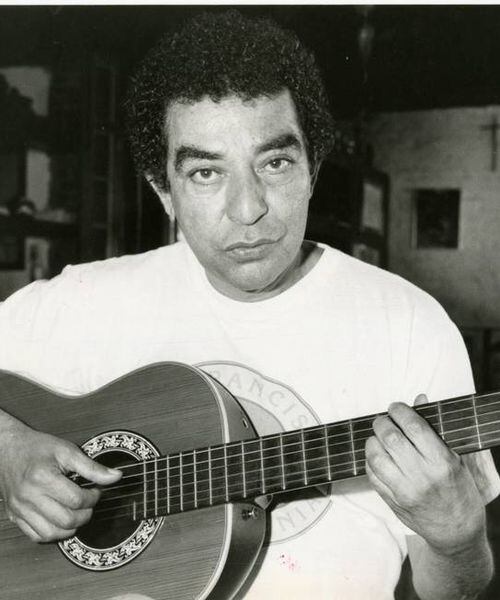 Foto em preto e branco. Músico João Nogueira sentado segurando seu violão