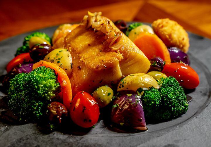 No centro de um prato raso cinza, estão servidas duas postas de bacalhau com brócolis, tomates, batatas e cebolas. O prato está sobre uma mesa de madeira.