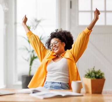 Joyful african american business woman freelancer   smiling and rejoices in victory while sitting at desk   and working at laptop screen after finishing project  in home office