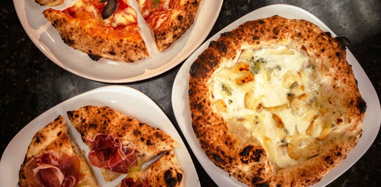 Em três pratos brancos, sobre uma mesa cinza, estão dispostas pizzas de diferentes sabores. Duas estão cortadas em quatro pedaços e uma está inteira. Foto: Paolo Ribeiro