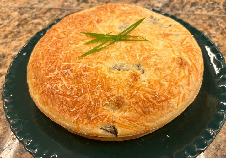 Sobre um prato de louça escura, uma torta de cogumelos com uma superfície com queijo parmesão ralado gratinado e com fatias de cebolinha para decorar por cima.