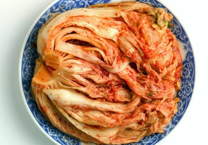 Prato branco com detalhes floridos azuis, com o alimento kimchi. O prato está sobre uma mesa branca.