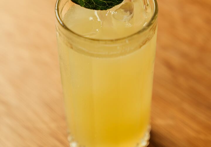 Um copo longo está sobre uma mesa de madeira e contém o drink amarelado, gelo e uma folha de hortelã por cima.