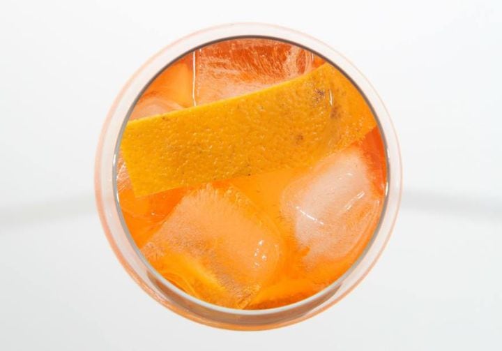 Copo visto de cima, com o drinque aperol spritz, com casca de laranja e cubos de gelo. O copo está sobre uma superfície branca.