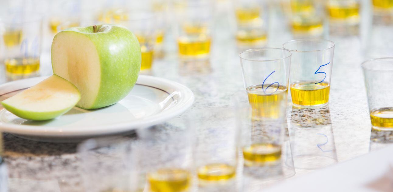 Copinhos de plástico transparente com um dedo de azeite em cada e prato de louça branca com uma maçã verde fatiada no centro. Foto: Tiago Queiroz