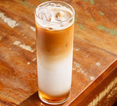 Café com leite servido com gelo em um copo longo de drink. O copo está servido em uma mesa rústica de madeira, com detalhes em branco e verde.