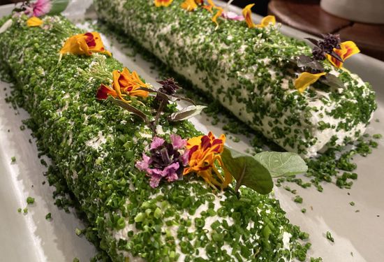 Em um prato retangular branco, estão dois cilindros de queijo lado a lado. Ambos estão enfeitados com ervas verdes e flores comestíveis amarelas e roxas. Foto: Dani Luci/Divulgação