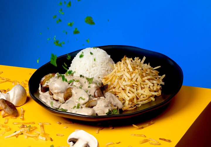 Strogonoff de plantas, servido com arroz e batata palha, em um prato preto sobre mesa de tampo amarelo e fundo azul. Há um pouco da guarnição e pedaços de cogumelos esparramados ao lado.