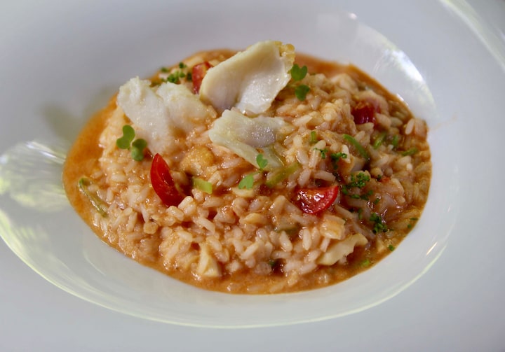 Um arroz de bacalhau, com tomate, lascas do peixe no topo e caldo na base, está servido em um prato branco, fundo e com bordas largas.
