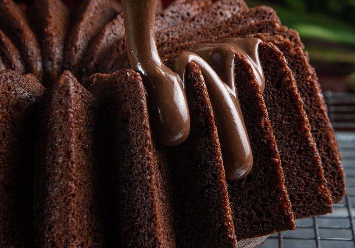 Sobre uma superfície de metal, há um bolo de chocolate. Uma generosa calda de chocolate, que está dentro de um jarro de vidro, está sendo derramada por cima do bolo.