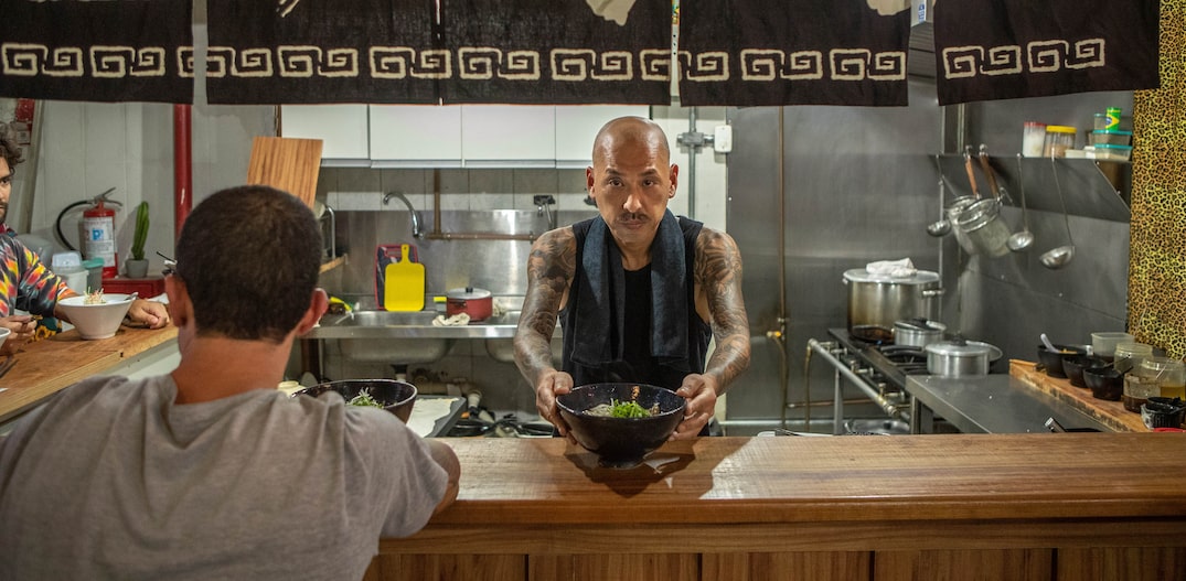 Na imagem, o japonês tatuado oferece uma tigela de lámen em direção à câmera.  Ao lado esquerdo, um cliente vestindo uma camiseta cinza degusta o prato. Foto: DANIEL TEIXEIRA