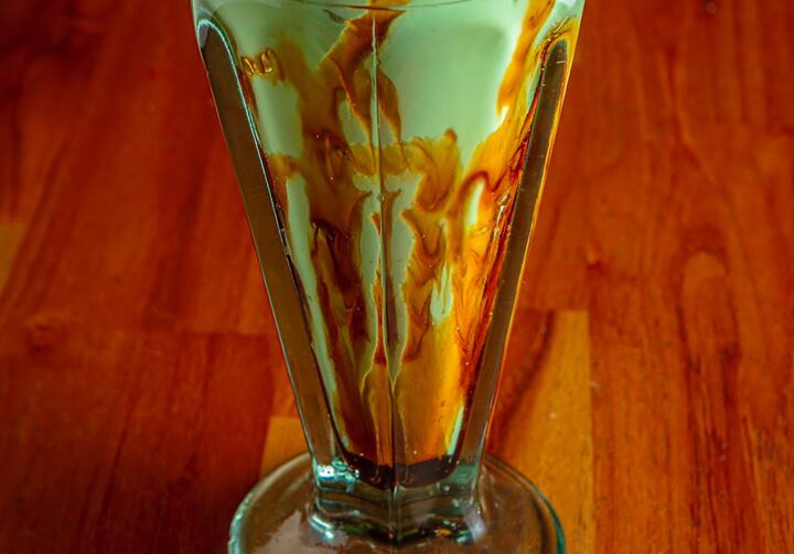 Em foco, uma taça de  milkshake de pistache com calda de caramelo, sobre uma mesa de madeira e com uma parede de madeira ao fundo.