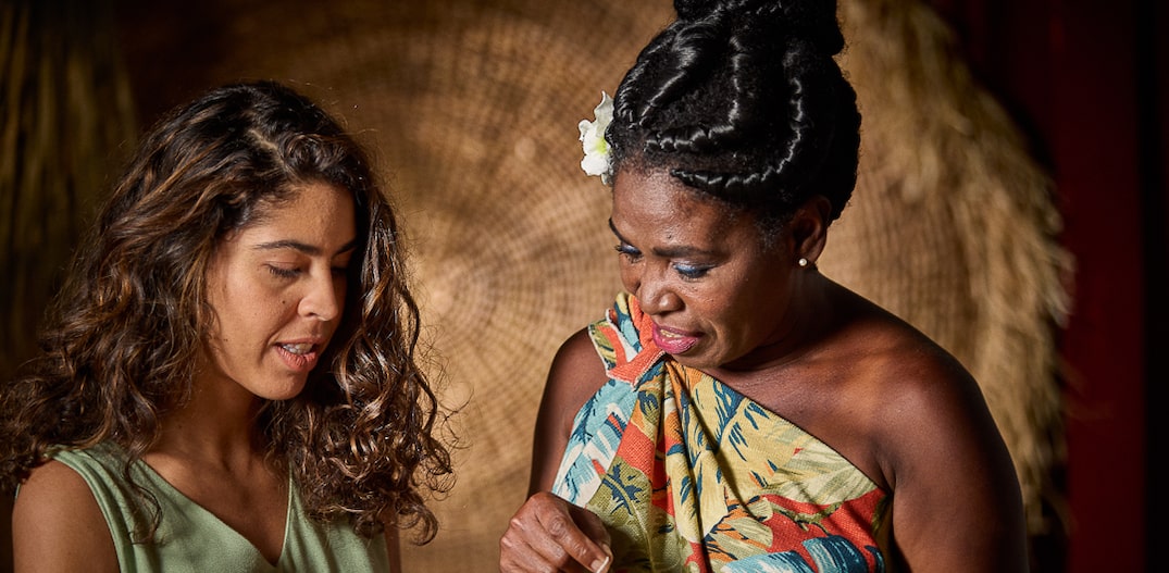 Duas mulheres negras manipulam alimento embrulhado em uma folha. elas vestem, respectivamente vestidos verde e estampado. Foto: Raphael Criscuolo