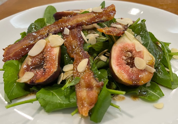 Em um prato branco, uma salada de rúcula com figos é exposta acompanhando lindas fatias de bacon e muitas lâminas de amêndoa.