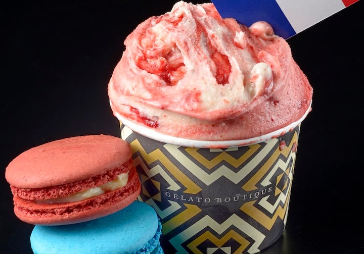 Dois macarons - um vermelho e um azul - estão ao lado de um copinho decorado com formas geométricas bege, branco e preto com um sorvete de tom avermelhado dentro. Uma bandeirinha da França está espetada no topo do sorvete. O fundo é preto