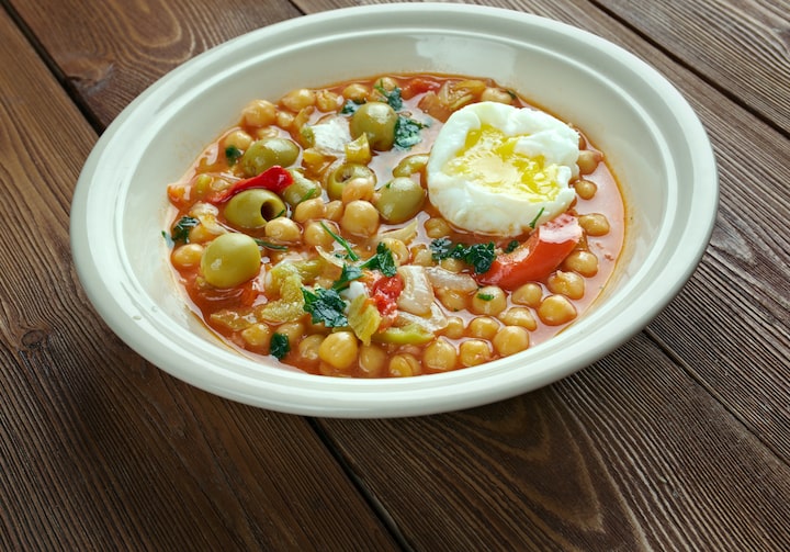 Em um prato branco e fundo está servido o Lablabi, que é uma comida que tem como base o grão de bico e outras coisas como ovos e azeitona, tudo fica em um molho vermelho. O prato está sobre uma mesa de madeira.