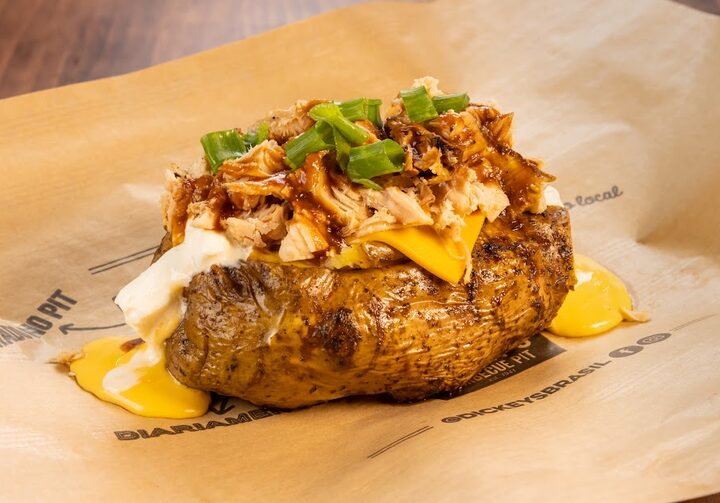 Sobre um papel pardo, está uma batata assada recheada com frango desfiado, cream cheese e queijo, enfeitado com cebolinhas e molho barbecue.