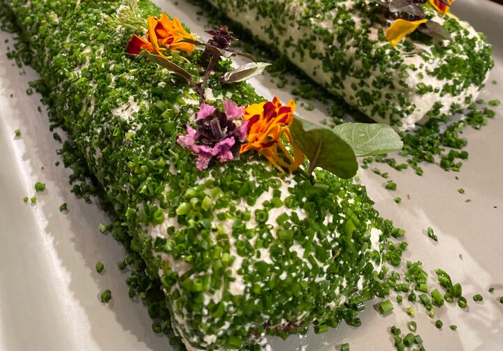 Em um prato retangular branco, estão dois cilindros de queijo lado a lado. Ambos estão enfeitados com ervas verdes e flores comestíveis amarelas e roxas.