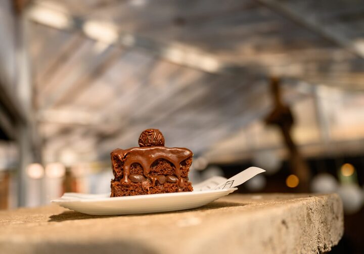Prato branco quadrado com pedaço de bolo de chocolate com um brigadeiro no topo. O prato está sobre uma mesa de concreto. Fundo desfocado.