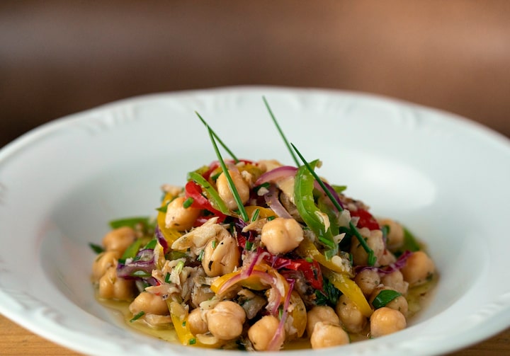 Salada de grão de bico e bacalhau, servida em um prato branco fundo, com bordas largas. As bordas do prato têm detalhes em relevo.