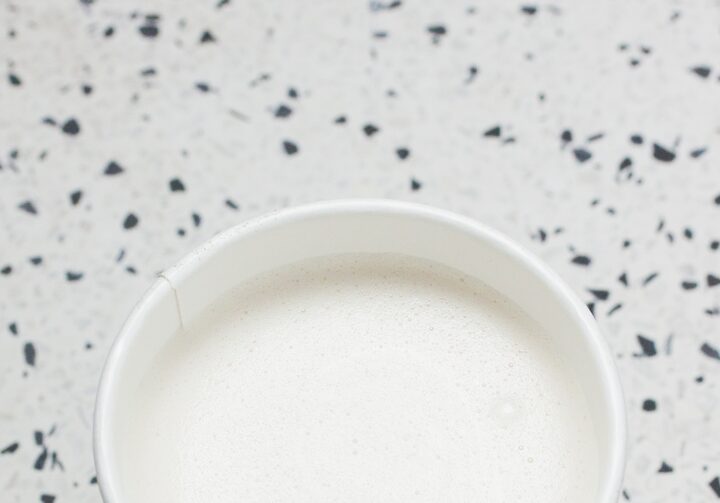 Copo branco, visto de cima, com espuma branca de leite, está sobre uma superfície de granito cinza, branco e preto. O copo de papelão tem o nome  do restaurante grafado em preto.