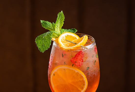 No canto de uma mesa de madeira está uma taça alta contendo o drink aperol rosé, com fatias de laranja, morangos e um buquê de hortelã enfeitando a bebida. Foto: Gustavo Pitta