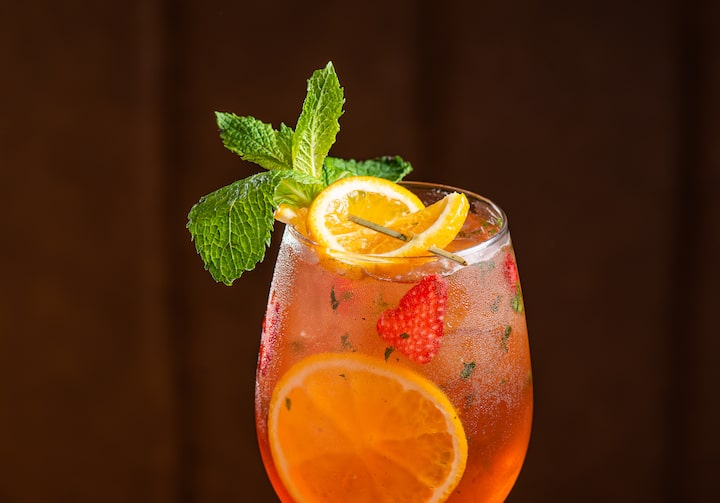 No canto de uma mesa de madeira está uma taça alta contendo o drink aperol rosé, com fatias de laranja, morangos e um buquê de hortelã enfeitando a bebida.