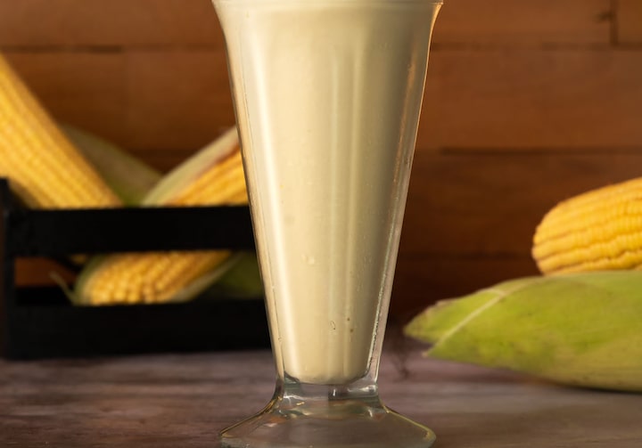 Em foco, uma taça de milkshake de milho, disposta sobre uma mesa de madeira com espigas de milho ao fundo.