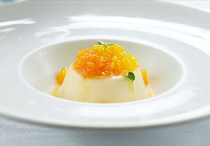 Prato fundo branco com um pequeno pudim branco - a panna cotta - coberto por laranja triturada, calda de laranja e duas pequenas folhas verdes. O prato está sobre uma mesa branca.