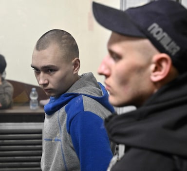 Russian soldier Vadim Shishimarin (C) looks on from the defendant's box at the opening of his trial on charge of War crimes for having killed a civilian, as his widow Kateryna Shelipova (L) reacts, in the Solomyansky district court in Kyiv on May 18, 2022. - The captured soldier is accused of killing a 62-year-old civilian -- allegedly on a bicycle -- near the village of Chupakhivka in the northeastern Ukraine Sumy region on February 28, in the first days of the Russian's offensive. Shishimarin pleaded guilty and is facing possible life imprisonment in Kyiv. (Photo by Genya SAVILOV / AFP)