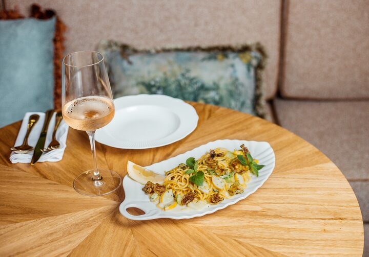 Spaghetti alla nerano servido em uma travessa branca que está ao lado de um prato raso,  uma taça de vinho branco, talheres e um guardanapo branco. As louças estão sobre uma mesa de madeira redonda. Há um sofá atrás, com duas almofadas azuis.