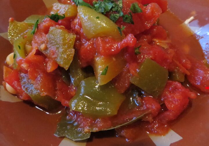 Em um prato de metal há pedaços de tomates e pimentões verdes decorados com salsa crespa. Os ingredientes escapam pelas bordas do prato e caem na superfície ocre com detalhes em branco.