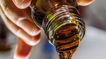Mão segurando um frasco de azeite, derramando o conteúdo em um copo transparente. Foto: Unsplash