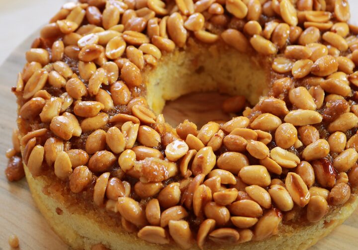 Um bolo de amendoim redondo e com um furo no meio está repleto de amendoins caramelizados por cima. O prato está servido sobre uma tábua de madeira.