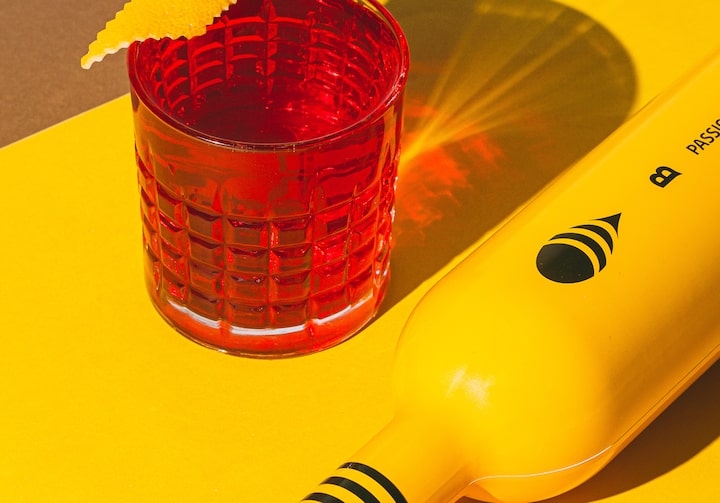 Drink na cor vermelha, em um copo baixo com um zest de laranja na borda. O copo está sobre uma mesa amarela. Ao lado dele, tem uma garrafa amarela com detalhes em preto deitada.