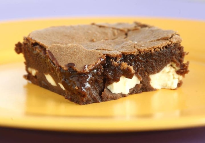 fatia quadrada de brownie de chocolate com predaços brancos de cream cheese no meio da massa, sobre um prato amarelo.