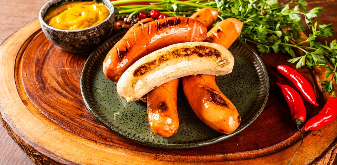 Salsichas alemãs grelhadas e dispostas sobre prato escuro e tábua de madeira, com talos de salsinha, pimenta malagueta e molho de mostarda ao lado. Foto: Alan/Berna