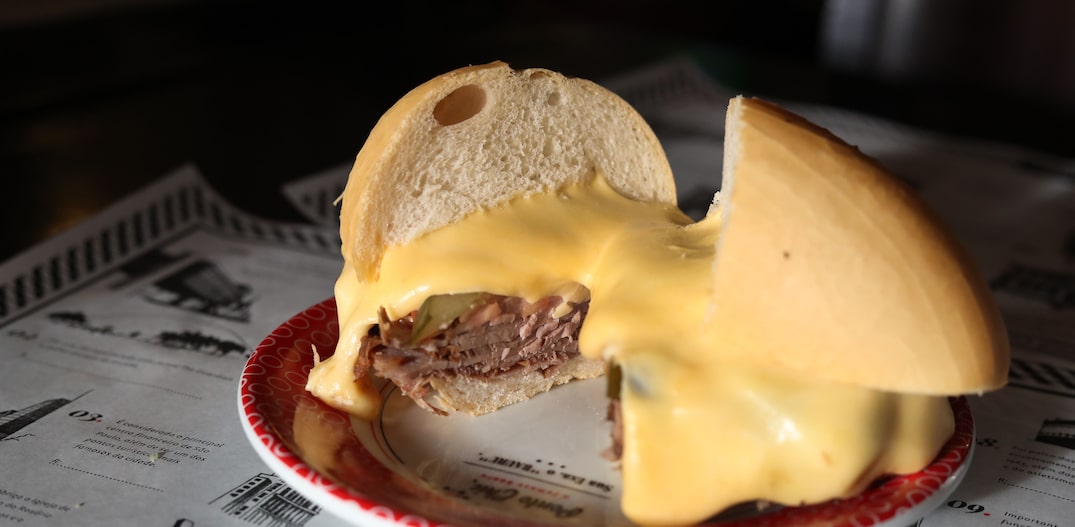 Em cima de um papel que lembra jornal, com as logos do Ponto Chic, está um prato com detalhes em vermelho e o sanduíche em cima, repartido e com o queijo derretendo. Foto: ALEX SILVA/ESTADAO