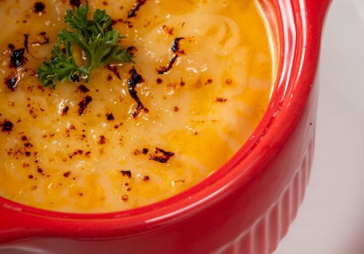 Em uma louça vermelha que imita uma pequena panela creuset, está uma porção de creme de cenoura com queijo e uma folhinha decorativa em cima