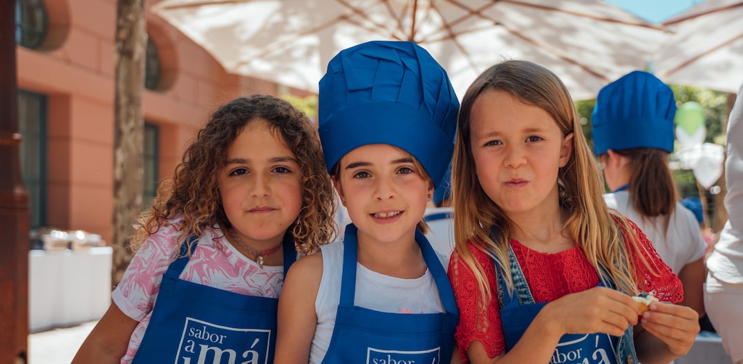 Três meninas com avental e chapéu de chefs de cozinha mostram um prato com pequenos doces. Foto: ©Anantara Hotels & Resorts / DIVULGAÇÂO