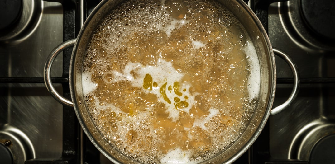 Macarrão cozinhando em água fervente, com pontos de azeite dispersos  sobre a superfície da água, em panela de aço inoxidável vista de cima. Foto: Roen/Adobe Stock