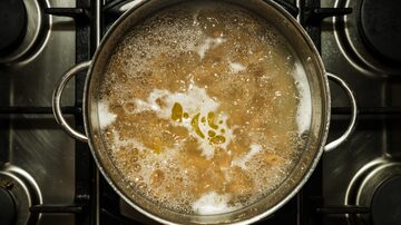 Macarrão cozinhando em água fervente, com pontos de azeite dispersos  sobre a superfície da água, em panela de aço inoxidável vista de cima. Foto: Roen/Adobe Stock