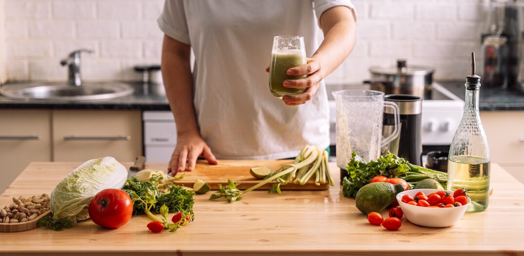 Lady in the kitchen holding a glass of smoothie in front of a wooden table on which there are fresh vegetables. Foto: Anastassiya | Adobe Stock