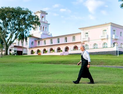 Homem branco com camisa branca e avental preto caminha sobre o gramado; ao fundo a fachada do Hotel das Cataratas, inteiramente pintado de cor de rosa claro com detalhes em branco