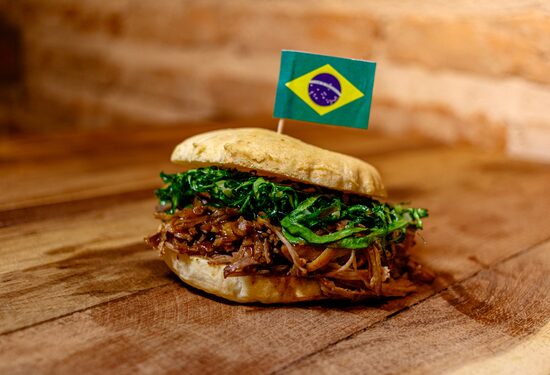 Em cima de uma mesa de madeira, está um pão de queijo grande recheado de pernil desfiado ao molho barbecue e tiras de rúcula. Em cima da fatia superior do pão, está fincada uma pequena bandeira do Brasil. Foto: Rodrigo Cafaro