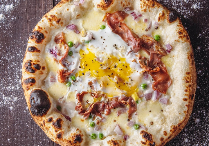 Pizza artesanal com queijo, molho bechamel, ervilhas cozidas, presunto de Parma e um ovo estalado ao meio. Isso, sobre uma mesa de madeira polvilhada com farinha de trigo.