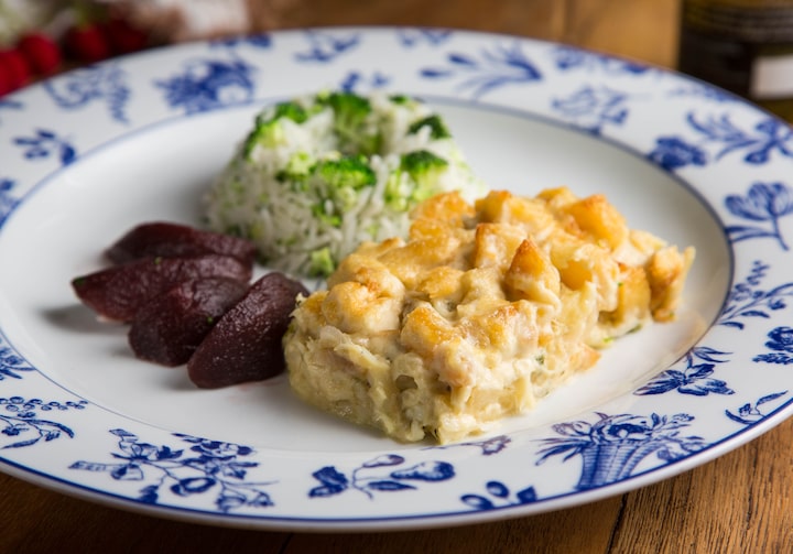 Em um prato branco, com desenho de flores em azul nas bordas, está servido o bacalhau com natas - um creme dourado - e peras ao vinho. Completando o prato, há uma porção de arroz com brócolis.