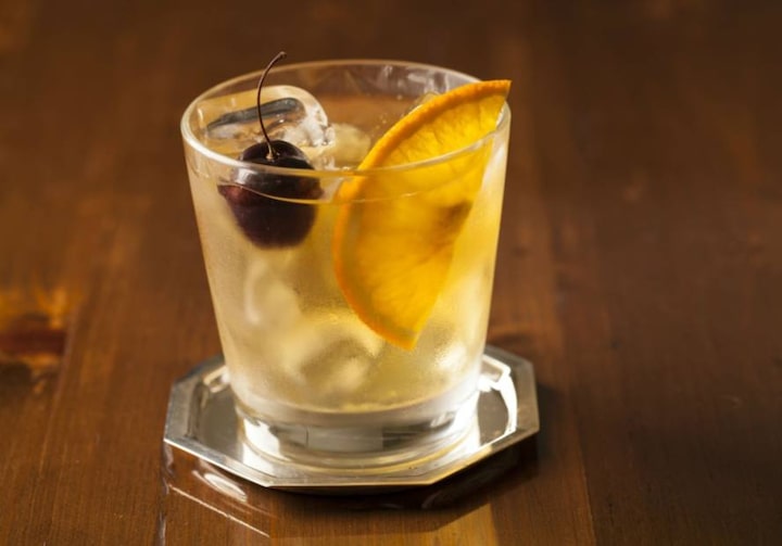 Mini prato de alumínio com copo transparente. O copo leva o drinque Old Fashioned, uma cereja e uma rodela de laranja. O copo está sob uma mesa de madeira marrom.