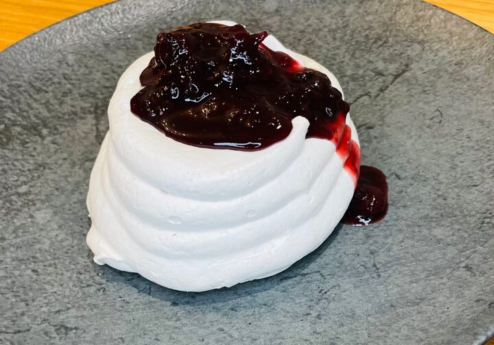 Doce branco em formato redondo com calda de frutas vermelhas no topo. O doce está em um prato cinza, sobre uma mesa de madeira.